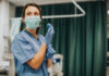 Nurse wearing scrubs and mask putting on gloves.