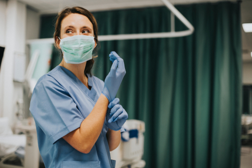 Nurse wearing scrubs and mask putting on gloves.