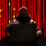 Hooded figure using laptop, China flag background with code.