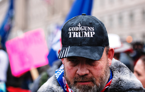 Man wearing God Guns Trump hat in a crowd.