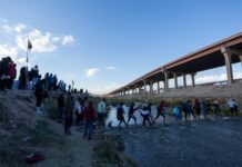 People crossing a shallow river by a bridge.