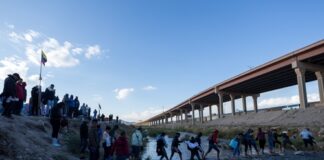 People crossing a shallow river by a bridge.