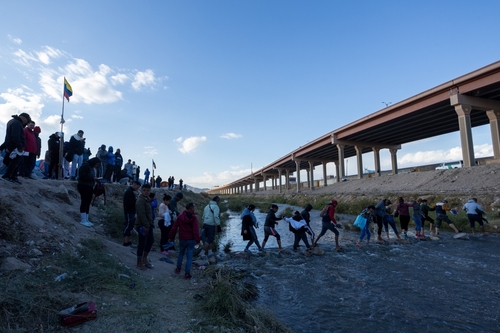 People crossing a shallow river by a bridge.