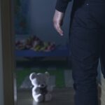 Man standing near a teddy bear in dim room.