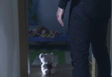 Man standing near a teddy bear in dim room.