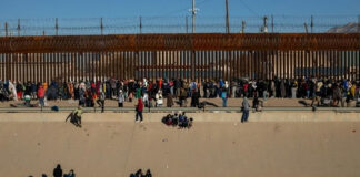 Large group of people gathered along a tall border fence near a concrete channel