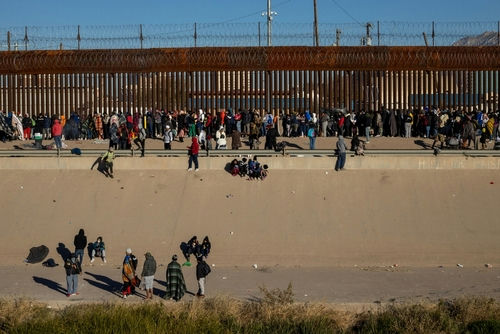 Large group of people gathered along a tall border fence near a concrete channel