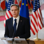 Trump’s Iran Deal SHOCKER—Strait of Hormuz to Reopen? Man in suit speaking at podium with international flags behind