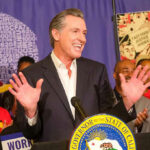 DOJ Firestorm Becomes Newsom’s ATM Man speaking at podium with smiling audience behind.