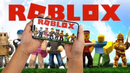 Hands holding a smartphone displaying the Roblox game with characters in the background