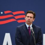 DeSantis Fumes Over COVID Insanity Ruling A public figure standing at a microphone with a serious expression against a backdrop featuring the USA flag