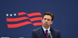 A public figure standing at a microphone with a serious expression against a backdrop featuring the USA flag