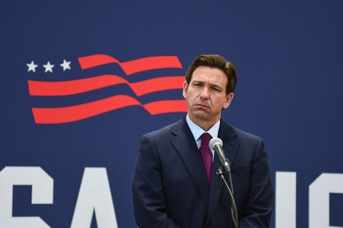 A public figure standing at a microphone with a serious expression against a backdrop featuring the USA flag