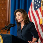 Patriot Millionaires? Hochul’s Desperate Pitch Woman speaking at podium with American flag backdrop.