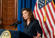 Patriot Millionaires? Hochul’s Desperate Pitch Woman speaking at podium with American flag backdrop.