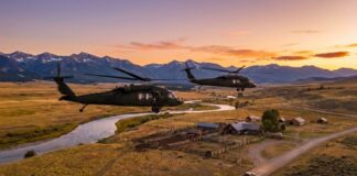 Two military helicopters flying over a rural landscape at sunset