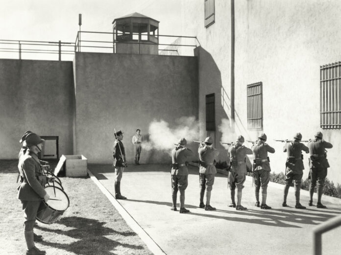 Facing,The,Firing,Squad Empty prison cell block with barred doors and walkway.