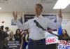 man in white shirt speaking at a rally with supporters and signs behind him