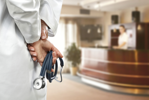 Your,Consultant,Of,Medicine,And,Slim,Woman,Hands A doctor holding a stethoscope behind their back in a hospital reception area
