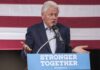 Epstein Entanglement Snags Clinton Insider man speaking at a podium with an American flag backdrop