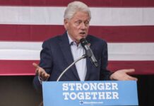 Epstein Entanglement Snags Clinton Insider man speaking at a podium with an American flag backdrop