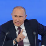 A political leader speaking at a press conference with a serious expression